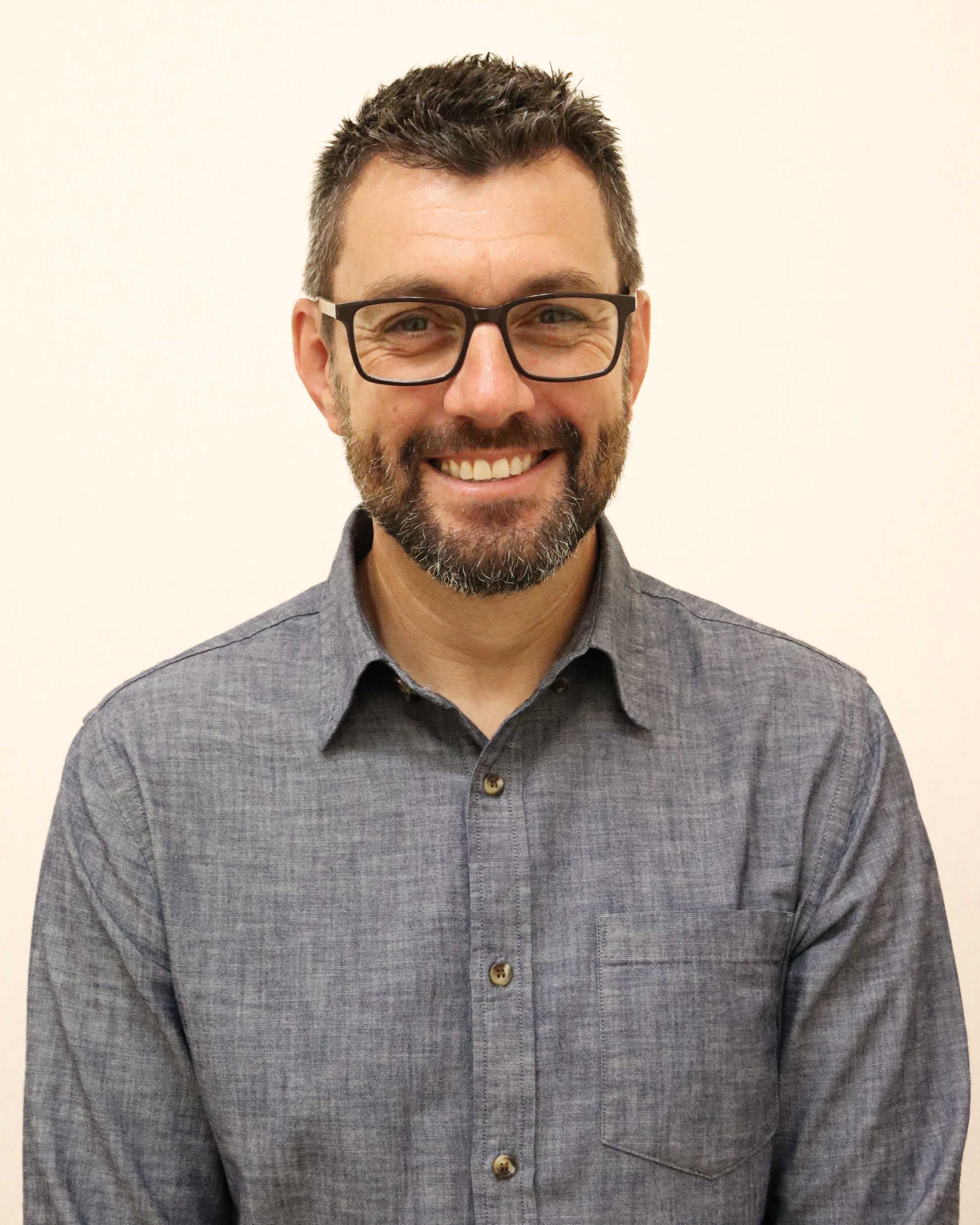 A photo of a man in a denim blue shirt, smiling at the camera